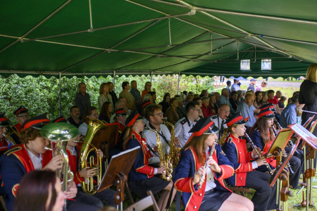 Grupa muzyków w czerwonych mundurach gra pod zielonym namiotem. Na widowni ludzie w tle. Instrumenty dęte i nuty widoczne.