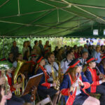 Grupa muzyków w czerwonych mundurach gra pod zielonym namiotem. Na widowni ludzie w tle. Instrumenty dęte i nuty widoczne.