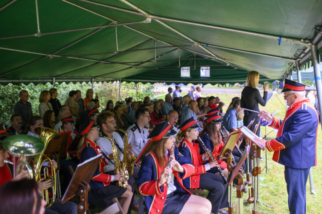Orkiestra w czerwono-niebieskich mundurach gra pod zielonym namiotem. W tle widoczna publiczność. Kobieta śpiewa przy mikrofonie.