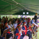 Orkiestra w czerwono-niebieskich mundurach gra pod zielonym namiotem. W tle widoczna publiczność. Kobieta śpiewa przy mikrofonie.