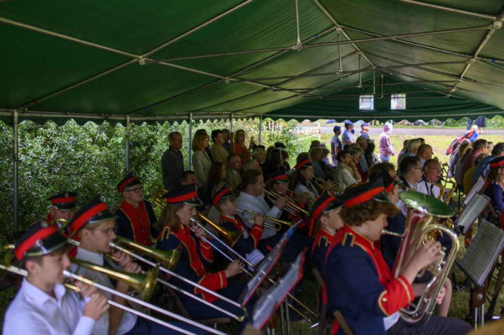 Orkiestra dęta w granatowych mundurach z czerwonymi akcentami gra pod zielonym namiotem. Publiczność siedzi w tle. Na zewnątrz zielone krzewy.