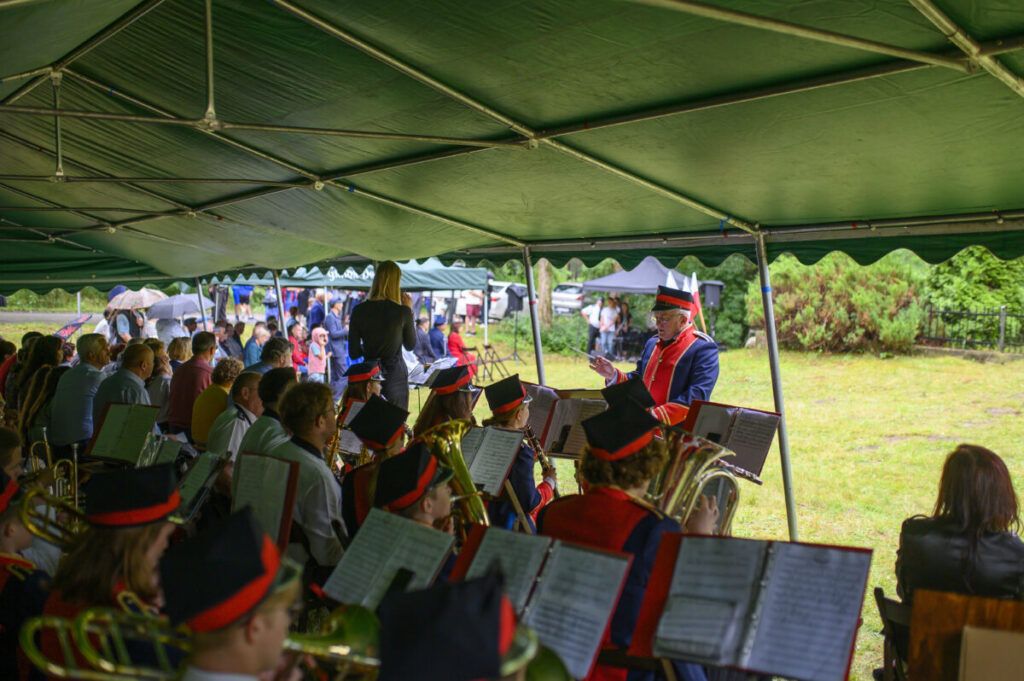 Orkiestra w kolorowych mundurach gra pod zielonym namiotem. Dyrygent w czerwonym płaszczu. Na tle zieleń i publiczność z parasolami.