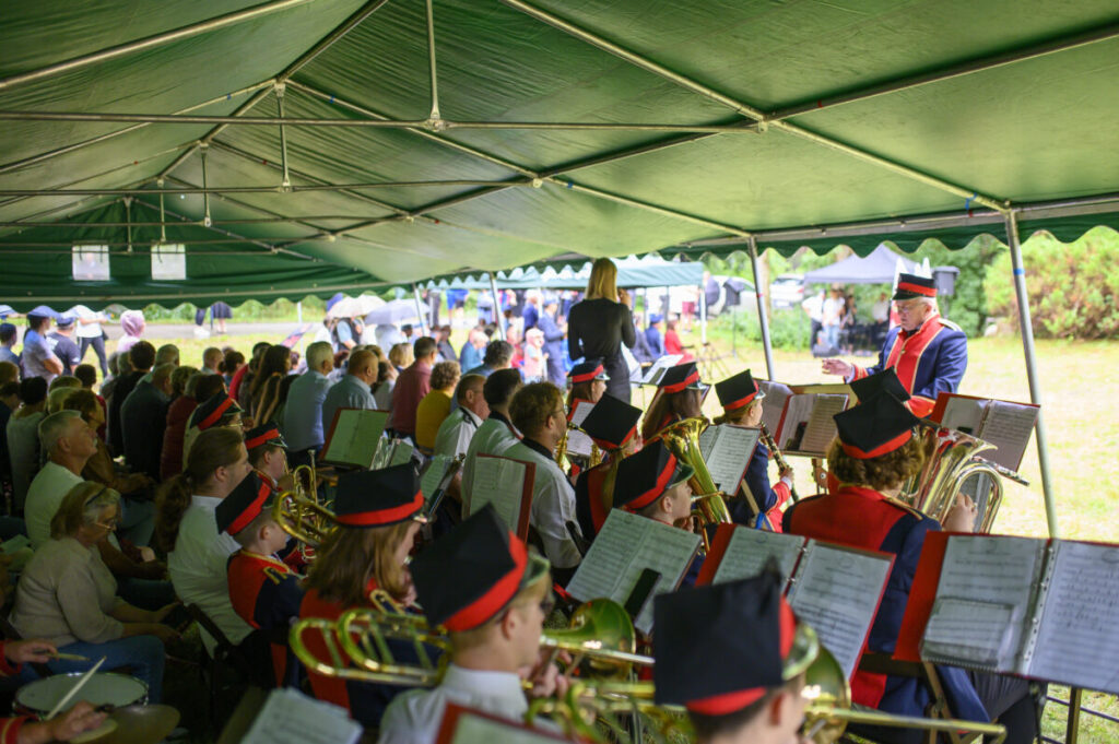 Orkiestra dęta w kolorowych mundurach gra pod zielonym namiotem. W tle widownia, na zewnątrz jasnozielona trawa. Dyrygent prowadzi muzyków.