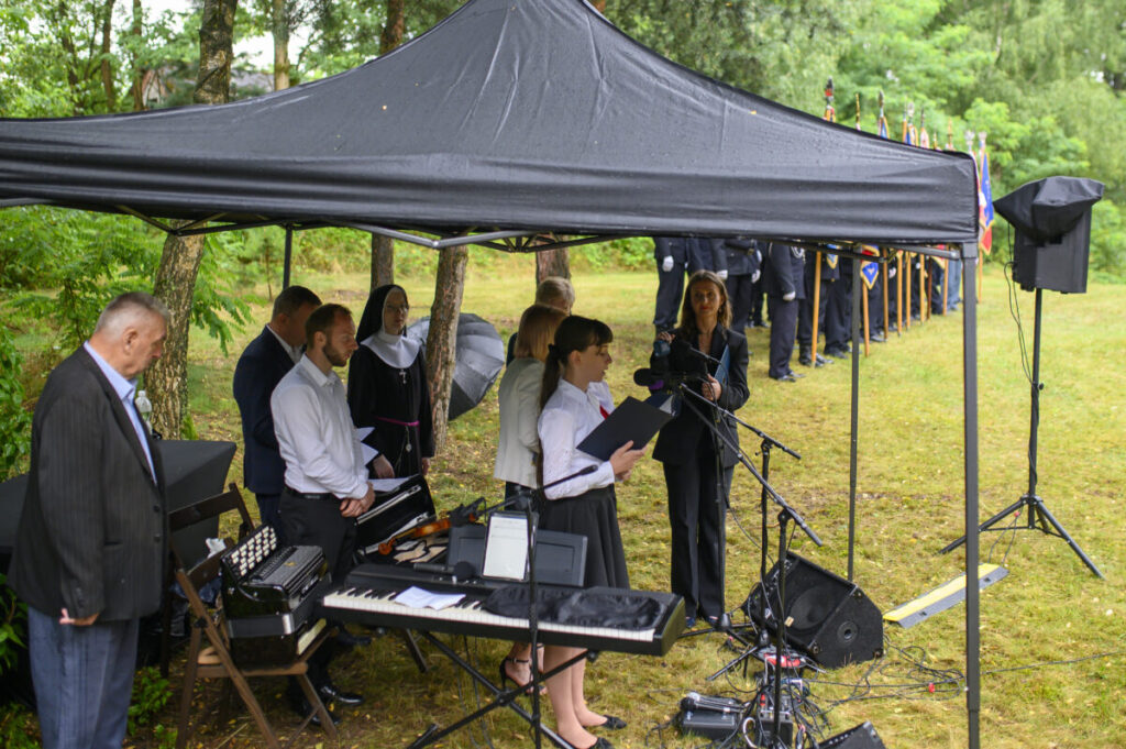 Grupa ludzi pod czarnym namiotem z mikrofonem i instrumentami, w tle zielone drzewa i stojąca straż honorowa.