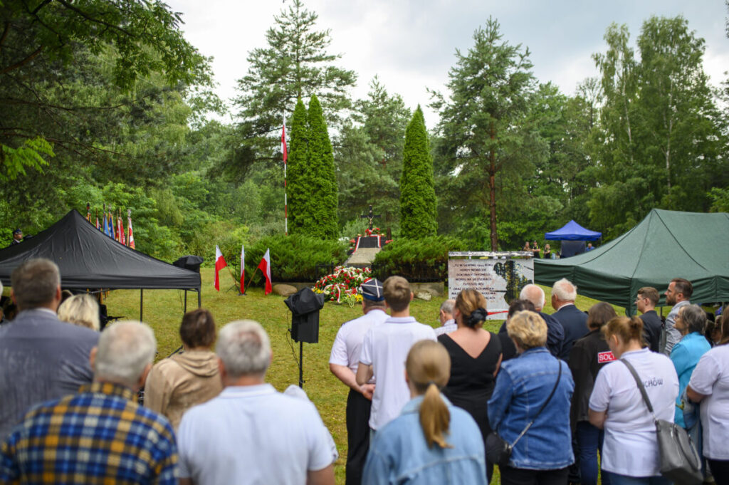 Na zdjęciu jest uroczystość plenerowa z flagami Polski, zielonymi drzewami i tłumem ludzi. Tło jest zielone, z drzewami i namiotami. Przed pomnikiem widać kwiaty i tablicę pamiątkową.