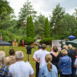 Na zdjęciu jest uroczystość plenerowa z flagami Polski, zielonymi drzewami i tłumem ludzi. Tło jest zielone, z drzewami i namiotami. Przed pomnikiem widać kwiaty i tablicę pamiątkową.