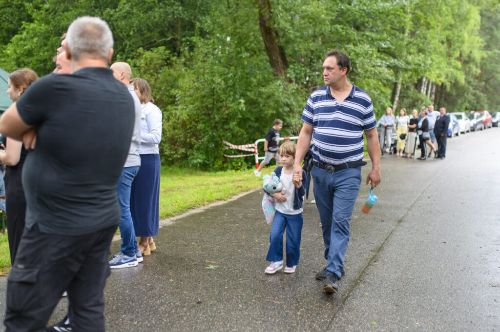 Na zdjęciu widzimy ludzi stojących w kolejce na mokrej drodze. Mężczyzna z dziewczynką trzymającą pluszaka idą obok. W tle zielone drzewa i zaparkowane samochody.