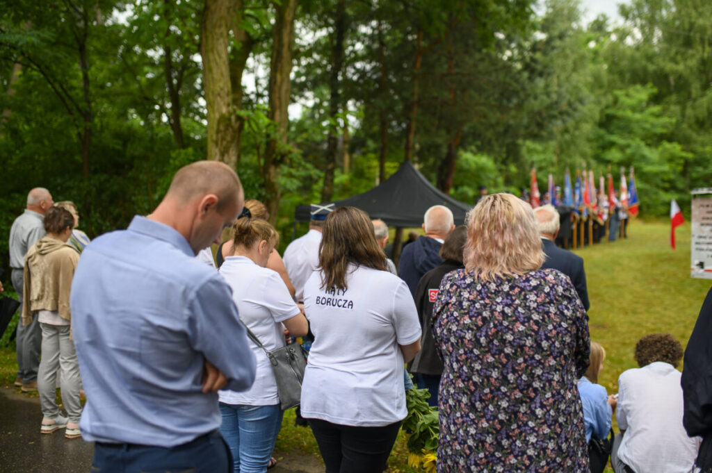 Grupa ludzi w parku uczestniczy w ceremonii. W tle flagi i namiot, otoczenie jest zielone. Dorośli i dzieci w odświętnych strojach.