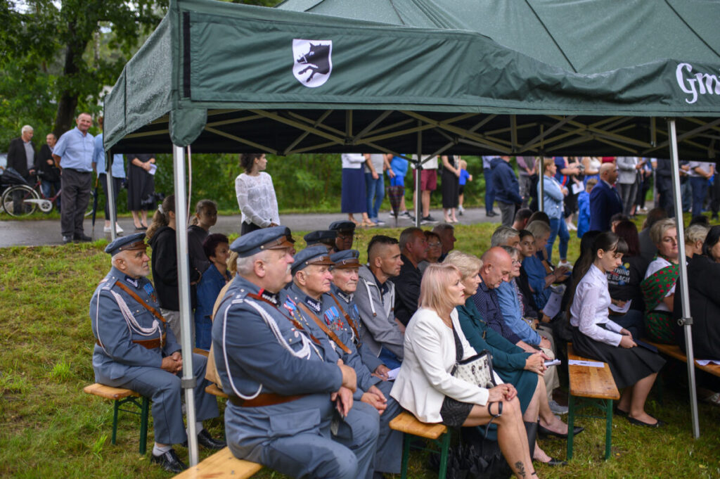 Grupa ludzi pod zielonym namiotem, część w mundurach, siedzi na ławkach. W tle widać widzów na trawie. Pogoda pochmurna.