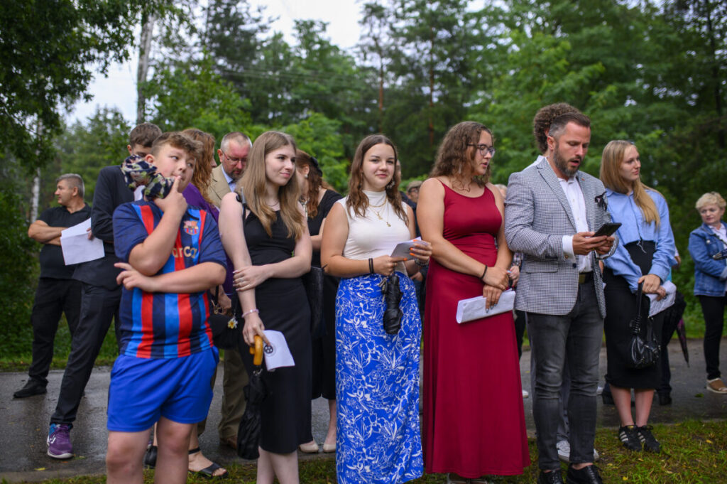 Grupa ludzi stoi na tle zielonych drzew, ubrana różnorodnie. Kobieta w czerwonej sukni wyróżnia się. Kilka osób trzyma kartki.