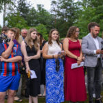 Grupa ludzi stoi na tle zielonych drzew, ubrana różnorodnie. Kobieta w czerwonej sukni wyróżnia się. Kilka osób trzyma kartki.