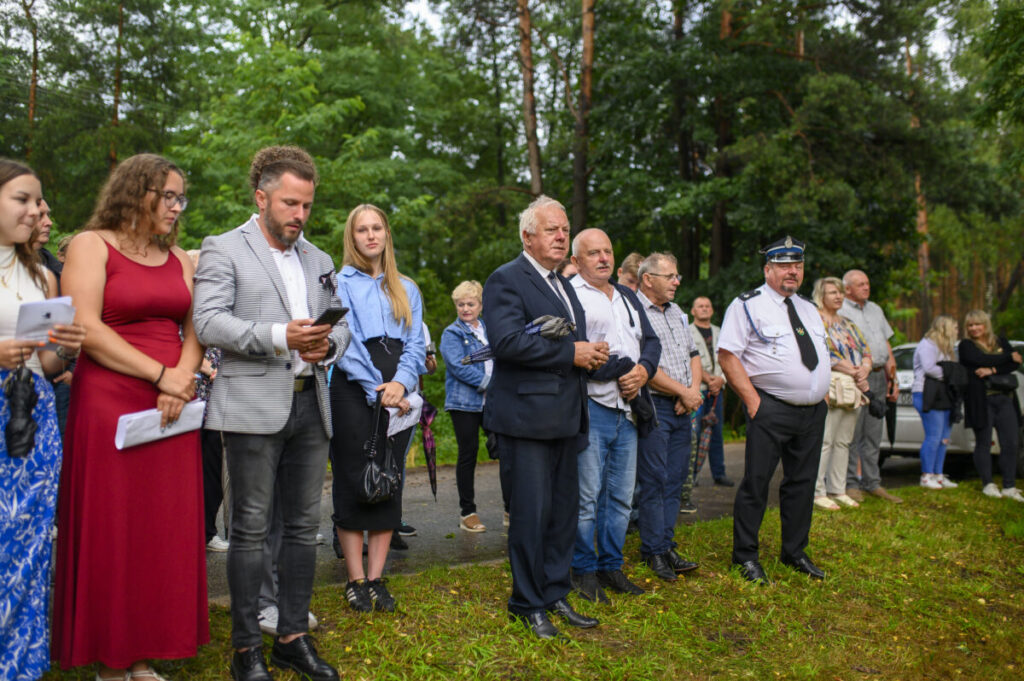 Grupa osób stoi na trawie w parkowym otoczeniu. Ubrani są elegancko, a w tle widać drzewa. Atmosfera jest formalna i spokojna.
