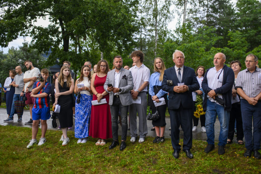 Grupa ludzi stoi na trawie, w eleganckich i casualowych strojach, na tle zieleni drzew. Część osób trzyma dokumenty. Pogoda jest pochmurna.