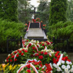 Na zdjęciu widoczny jest grób ozdobiony wieńcami z czerwonych i białych kwiatów, z krzyżem w tle. Tło to zielone drzewka i krzewy. Na wieńcach widoczne są biało-czerwone wstęgi.