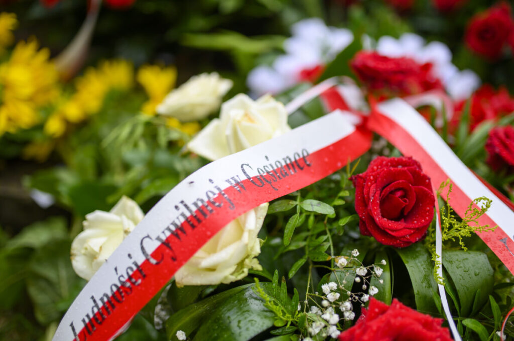 Na zdjęciu widać czerwone i białe róże ozdobione biało-czerwoną wstęgą z napisem. Tło stanowią zielone liście. Widoczne są też żółte kwiaty w tle.