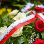 Na zdjęciu widać czerwone i białe róże ozdobione biało-czerwoną wstęgą z napisem. Tło stanowią zielone liście. Widoczne są też żółte kwiaty w tle.