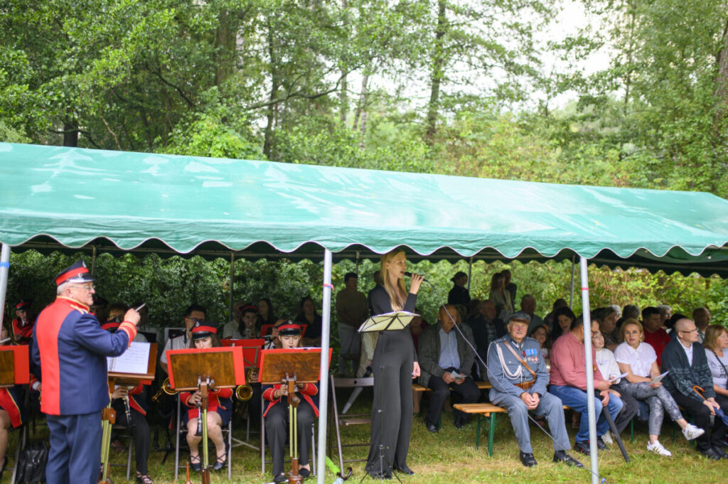 Grupa osób siedzi pod zielonym daszkiem. Na pierwszym planie muzyk dyryguje orkiestrą, kobieta śpiewa do mikrofonu. W tle widać drzewa.