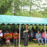 Grupa osób siedzi pod zielonym daszkiem. Na pierwszym planie muzyk dyryguje orkiestrą, kobieta śpiewa do mikrofonu. W tle widać drzewa.