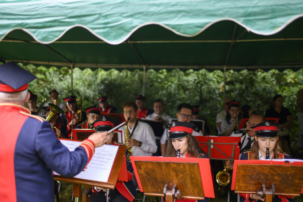 Orkiestra w czerwonych mundurach gra pod zielonym namiotem. Dyrygent kieruje zespołem. W tle zielone krzewy.