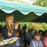 Kobieta w czarnym ubraniu śpiewa do mikrofonu. W tle zielony namiot i siedzący ludzie, w tym starszy mężczyzna w mundurze z medalami. Słoneczny dzień, natura w tle.