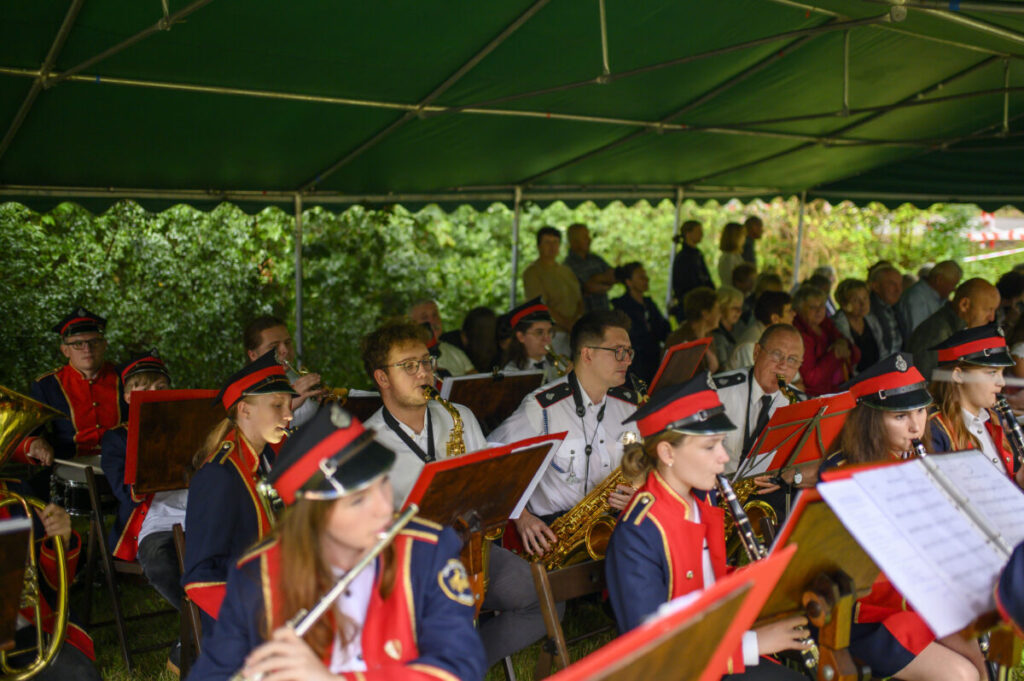 Orkiestra dęta gra pod zielonym namiotem. Członkowie noszą czerwono-granatowe mundury i czarne czapki. W tle widać widownię.