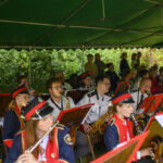 Orkiestra dęta gra pod zielonym namiotem. Członkowie noszą czerwono-granatowe mundury i czarne czapki. W tle widać widownię.