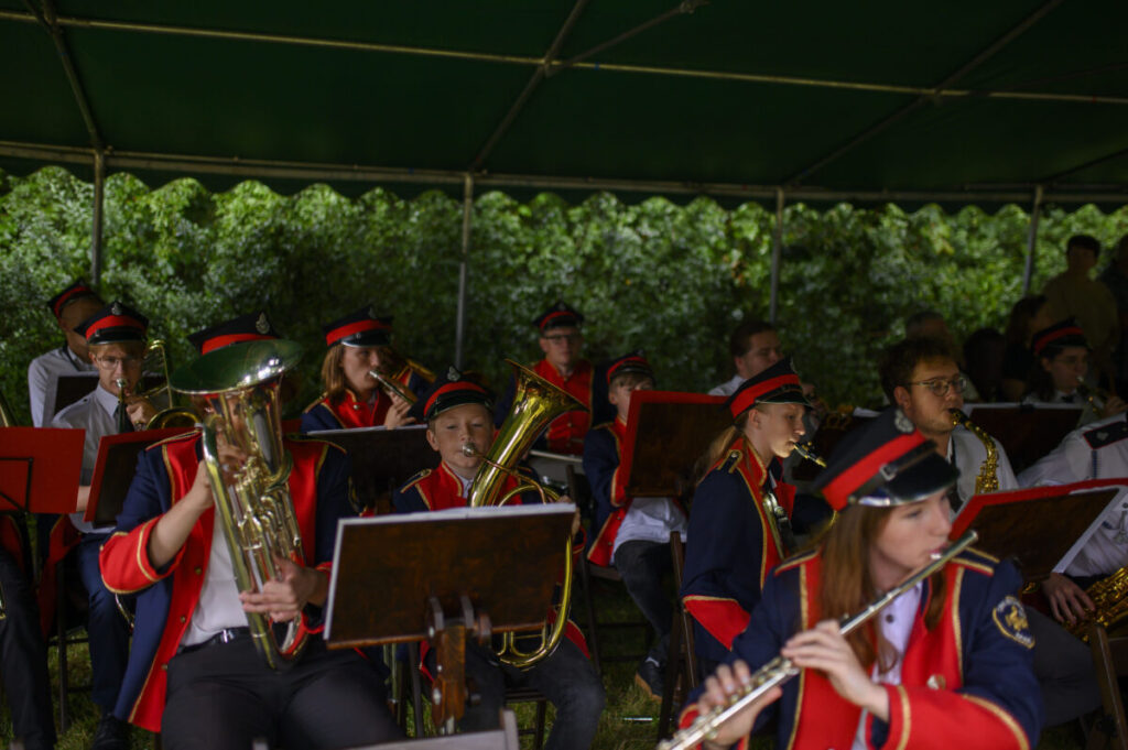 Orkiestra w czerwono-niebieskich mundurach gra pod zielonym namiotem. Tło to bujna zieleń. Instrumenty to głównie dęte i saksofony.