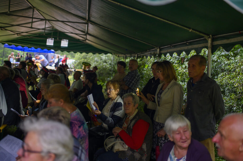 Grupa ludzi siedząca pod zielonym namiotem na tle krzewów. Osoby trzymają kartki z tekstem. Na dalszym planie niebieski daszek.
