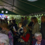 Grupa ludzi siedząca pod zielonym namiotem na tle krzewów. Osoby trzymają kartki z tekstem. Na dalszym planie niebieski daszek.