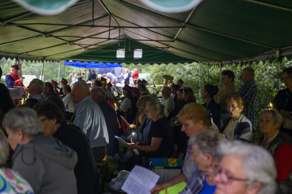 Ludzie siedzą pod zielonym namiotem podczas uroczystości. Kilka osób trzyma świece, a w tle są muzycy w strojach. Dzień jest pochmurny, a za namiotem widać zielone krzewy.