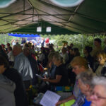 Ludzie siedzą pod zielonym namiotem podczas uroczystości. Kilka osób trzyma świece, a w tle są muzycy w strojach. Dzień jest pochmurny, a za namiotem widać zielone krzewy.