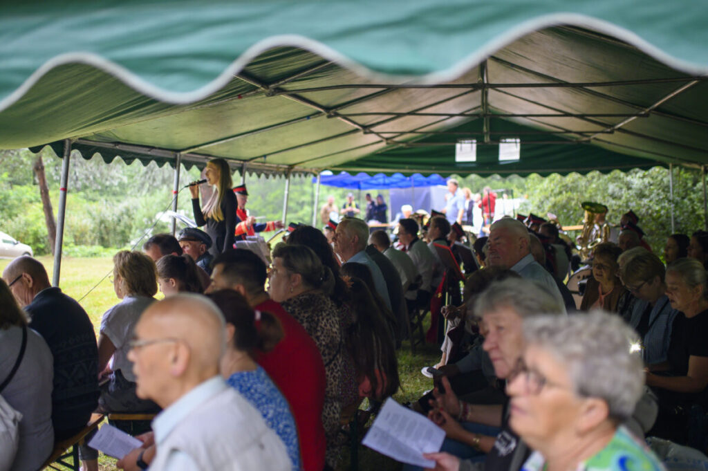 Ludzie siedzą pod zielonym namiotem na zewnątrz, słuchając mówcy. W tle drzewa i inne namioty. Atmosfera jest spokojna i zrelaksowana.