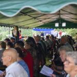 Ludzie siedzą pod zielonym namiotem na zewnątrz, słuchając mówcy. W tle drzewa i inne namioty. Atmosfera jest spokojna i zrelaksowana.