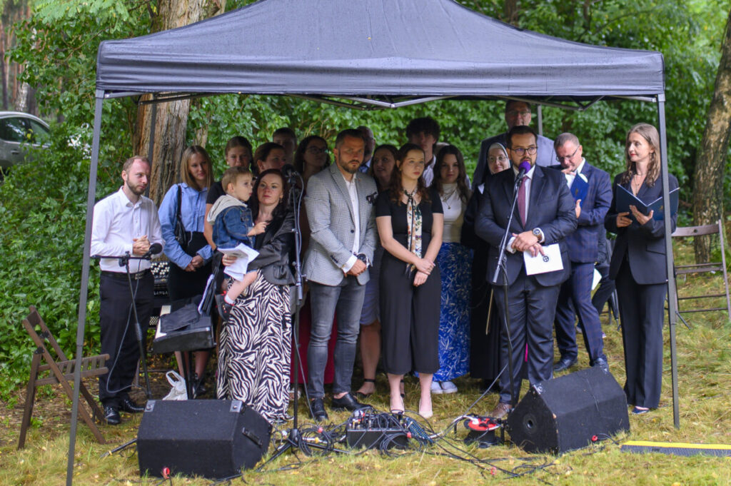 Grupa ludzi pod czarnym namiotem na tle zieleni. Osoby są elegancko ubrane, część trzyma mikrofony i dokumenty. Tło to drzewa i samochód.