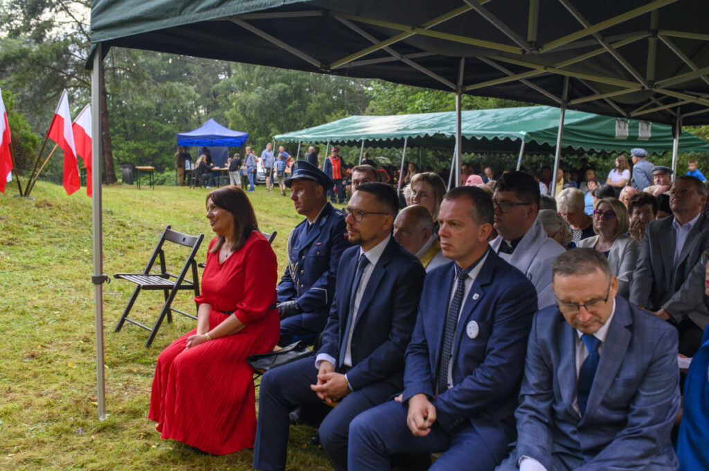 Festyn na trawie z widownią w garniturach, kobieta w czerwieni. Tła są zielone namioty i drzewa, flagi narodowe.