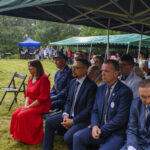 Festyn na trawie z widownią w garniturach, kobieta w czerwieni. Tła są zielone namioty i drzewa, flagi narodowe.