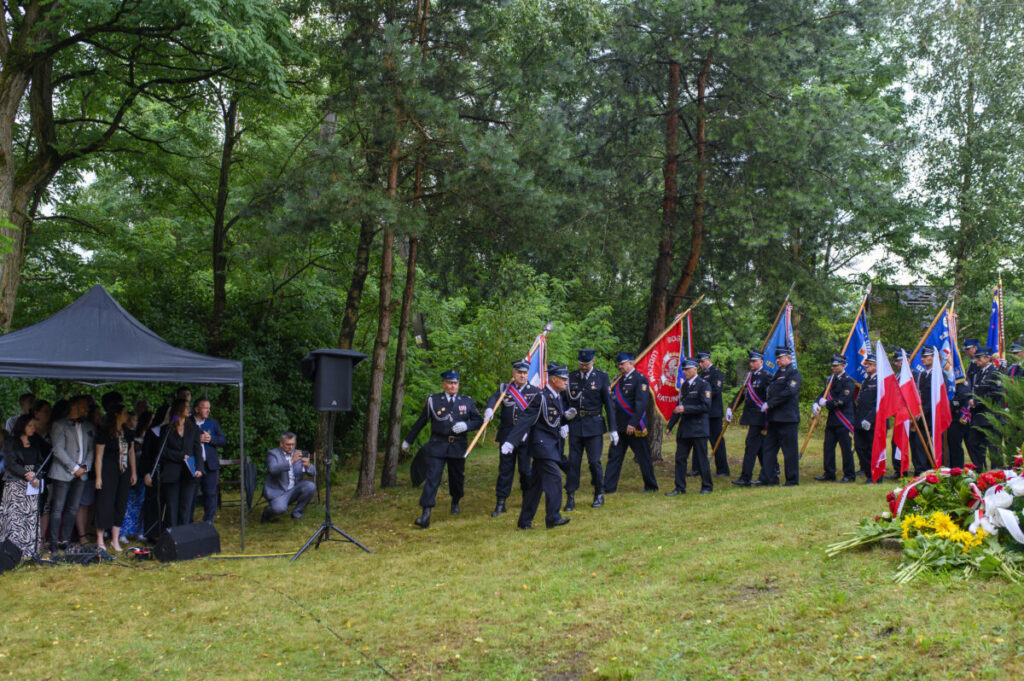 Grupa strażaków maszeruje z flagami po zielonym trawniku, otoczonym drzewami. Na lewo stoi namiot z widownią. Na trawie leżą kwiaty.