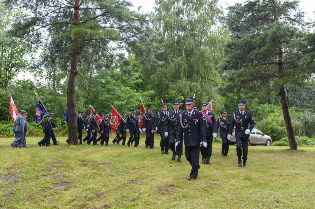 Grupa ludzi w mundurach maszeruje z flagami przez zielony teren. W tle drzewa, niebiesko-czarne ubrania. Spokój i porządek.