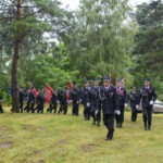 Grupa ludzi w mundurach maszeruje z flagami przez zielony teren. W tle drzewa, niebiesko-czarne ubrania. Spokój i porządek.