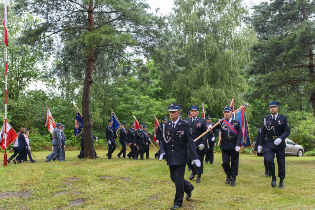 Grupa osób w mundurach maszeruje z flagami na tle zielonego parku. Drzewo i samochód w tle. Pogodny dzień.