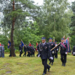 Grupa osób w mundurach maszeruje z flagami na tle zielonego parku. Drzewo i samochód w tle. Pogodny dzień.