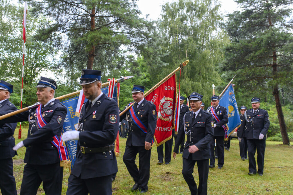 Na zdjęciu widać grupę strażaków w paradnych mundurach niosących sztandary. Tło stanowią zielone drzewa i trawa. Kolory dominujące to czerń mundurów oraz czerwony i niebieski sztandarów.
