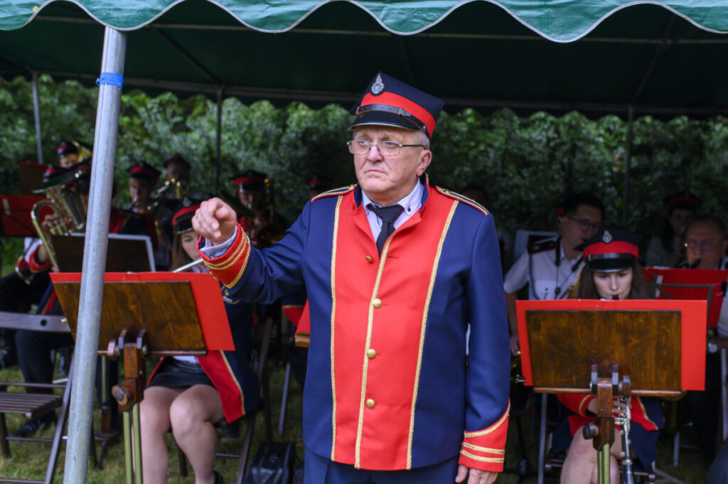 Dyrygent w kolorowym mundurze stoi przed orkiestrą. Czerwono-niebieski strój wyróżnia się na tle zieleni. Muzycy grają pod zielonym namiotem.