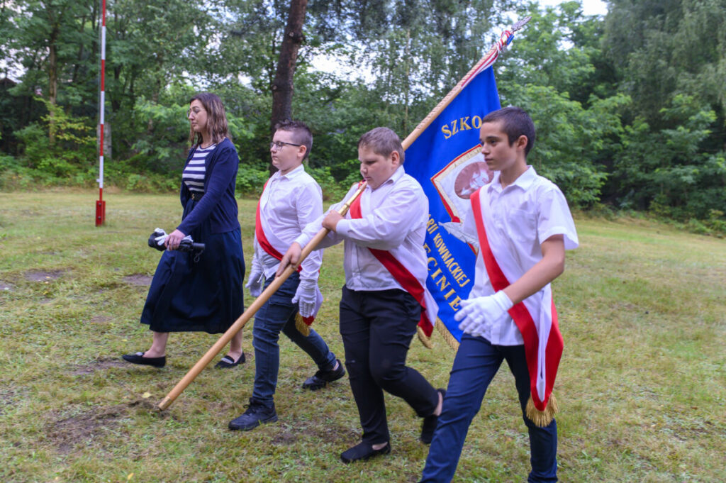 Na zdjęciu grupa młodych osób w białych koszulach niesie niebieską flagę. Tło stanowi zielona trawa i drzewa. Atmosfera jest uroczysta, a kolory żywe.