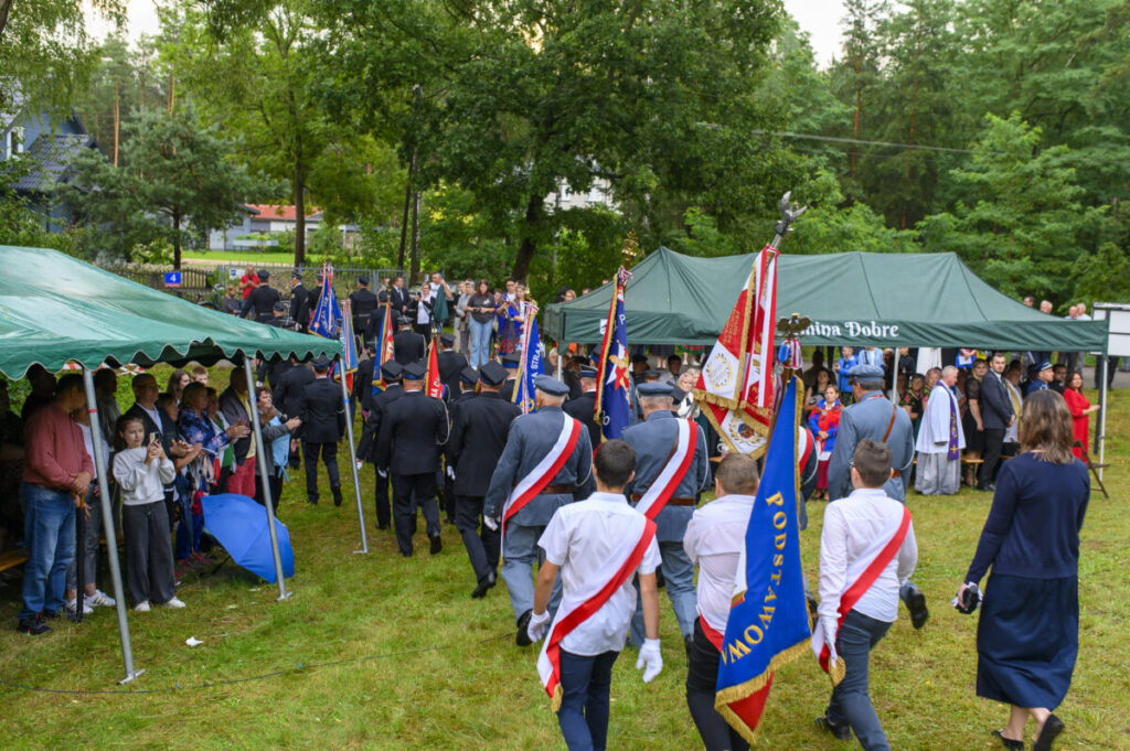 Na zdjęciu widać grupę ludzi w mundurach i odświętnych strojach z flagami. Uczestnicy idą przez trawnik pod zielonymi namiotami. W tle widoczne są drzewa i zebrani ludzie.