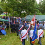 Na zdjęciu widać grupę ludzi w mundurach i odświętnych strojach z flagami. Uczestnicy idą przez trawnik pod zielonymi namiotami. W tle widoczne są drzewa i zebrani ludzie.