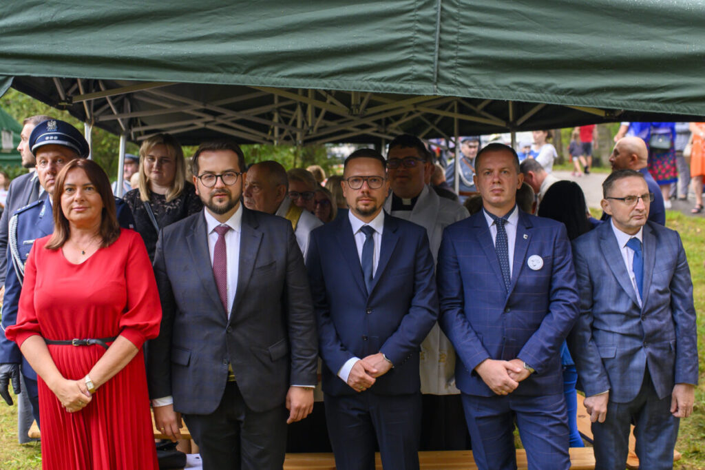 Pod zielonym namiotem stoi grupa elegancko ubranych osób. Mężczyźni w garniturach, kobieta w czerwonej sukience. W tle widać uczestników wydarzenia.