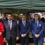 Grupa ludzi w eleganckich strojach stoi pod zielonym namiotem. Mężczyźni w garniturach, kobieta w czerwonej sukience. W tle widać osoby i zieleń.