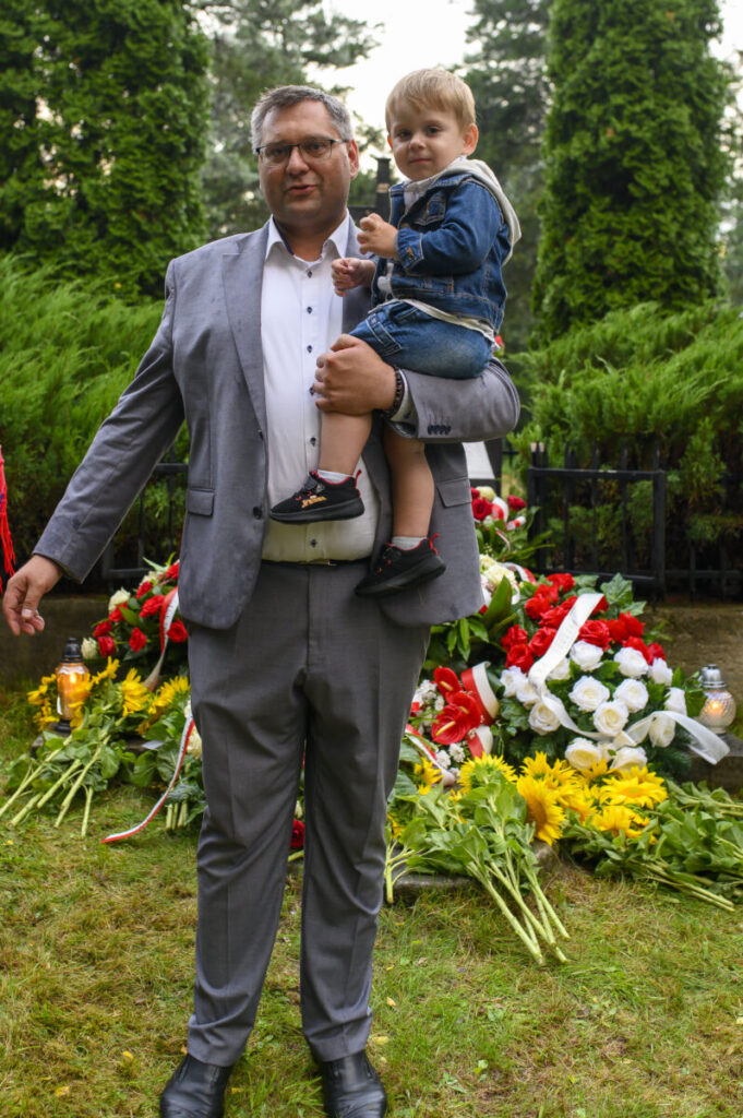 Mężczyzna w szarym garniturze trzyma dziecko na tle kolorowych kwiatów. Wokół zieleń i drzewa. Spokojne, naturalne otoczenie.