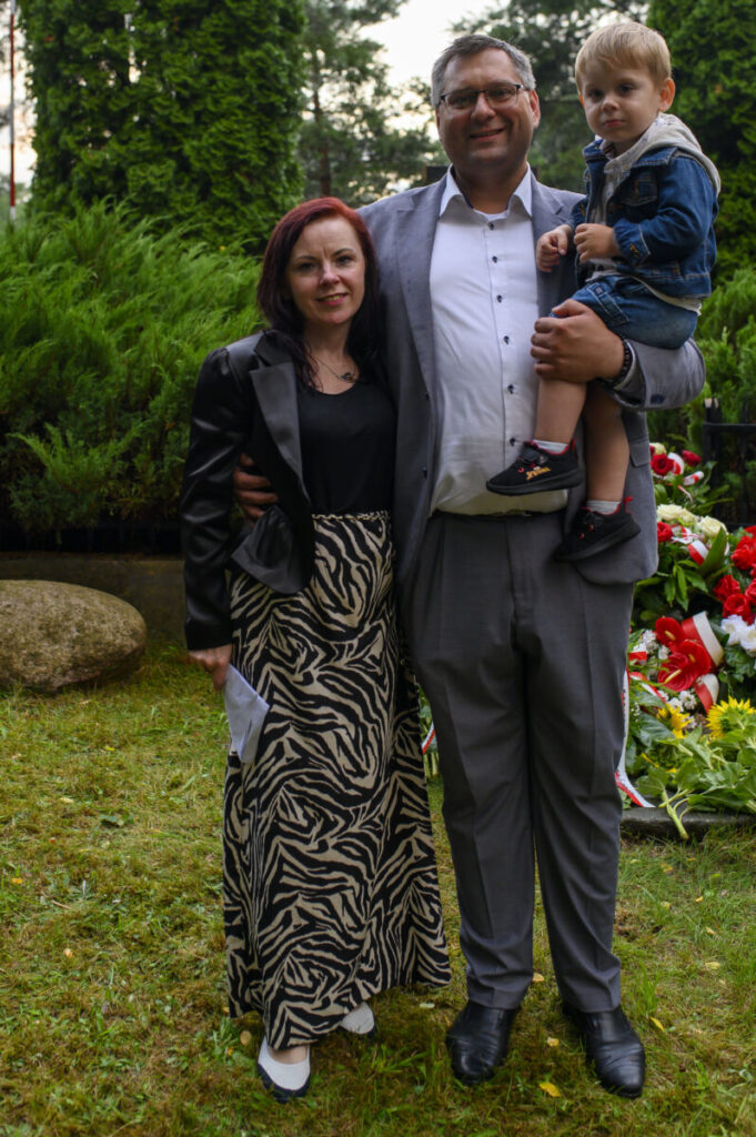 Grupa osób pozuje na trawie, w tle zieleń i kwiaty. Mężczyzna trzyma dziecko. Kobieta w czarno-białej spódnicy.